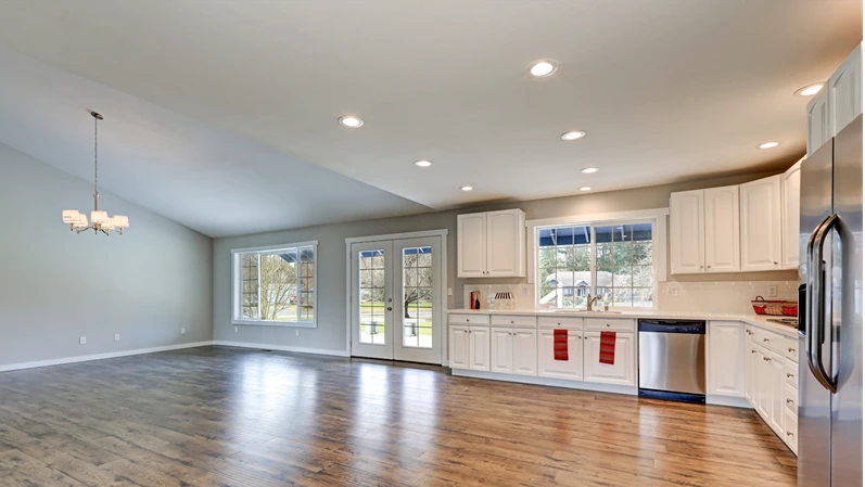 Spacious rambler home interior with vaulted ceiling over glossy laminate floor. Light filled kitchen room boasts white cabinets and stainless steel appliances. Northwest, USA