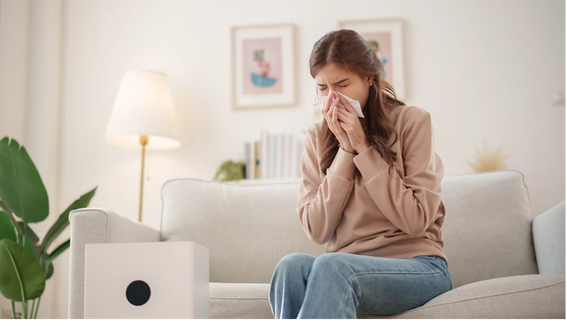 Young Asian woman sits on a couch, sneezing and holding a tissue to her nose while an air purifier operates nearby to improve air quality.