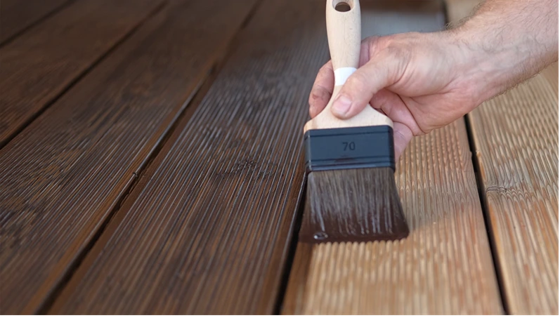 Brown oil for protection of wood by staining. Painting a larch for protecting and pests. Woodworker applying finish to terrace deck board. Repairman varnishing with brush wooden plank floor surface.