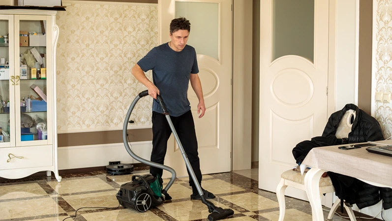 A man in a blue shirt pushes a green vacuum across the tiled floor, moving the vacuum cleaner from room to room