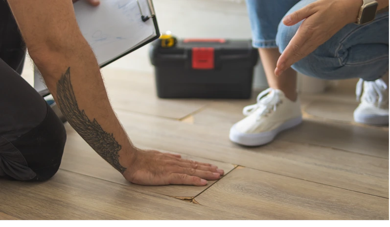 Contractor pointing at buckling laminate flooring, discussing home repair and renovation options with a homeowner, emphasizing the need for flooring replacement due to damage