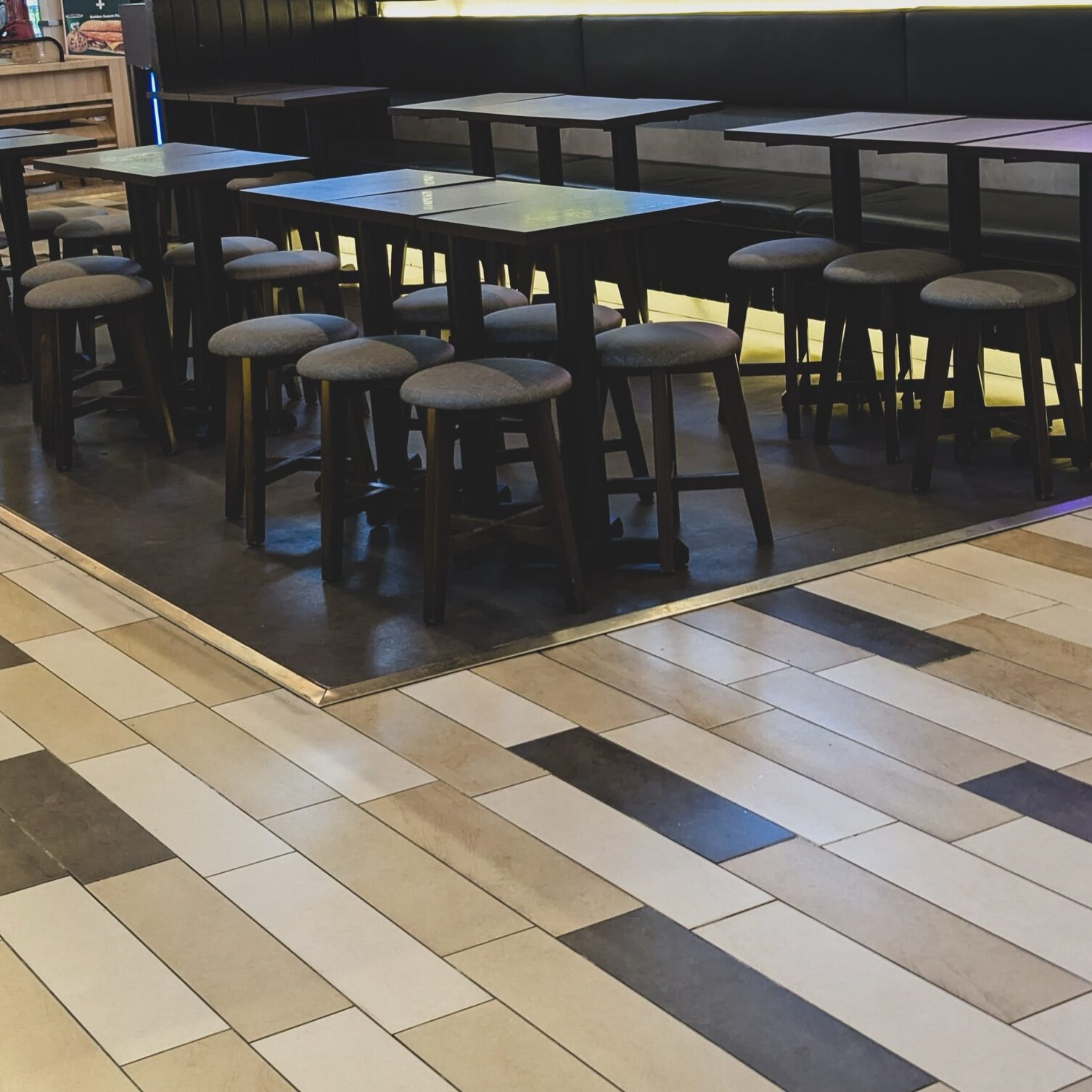 Interior of a modern cafeteria or restaurant featuring rows of long wooden tables with dark metal legs and round grey stools on a tiled floor.